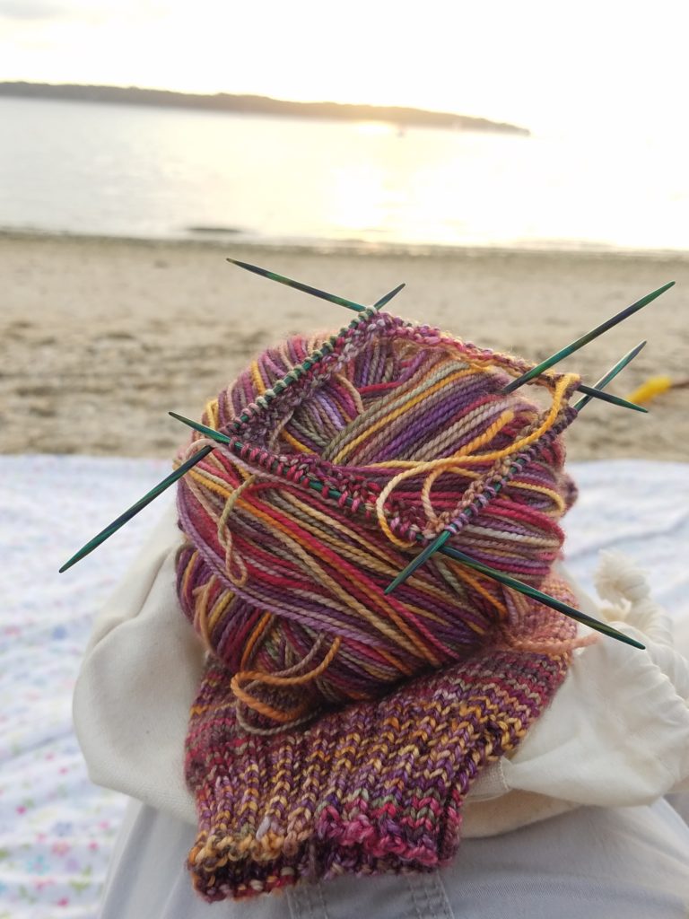 A newly cast-on sock lying on a beach blanket with the Long Island Sound at sunset in the background.