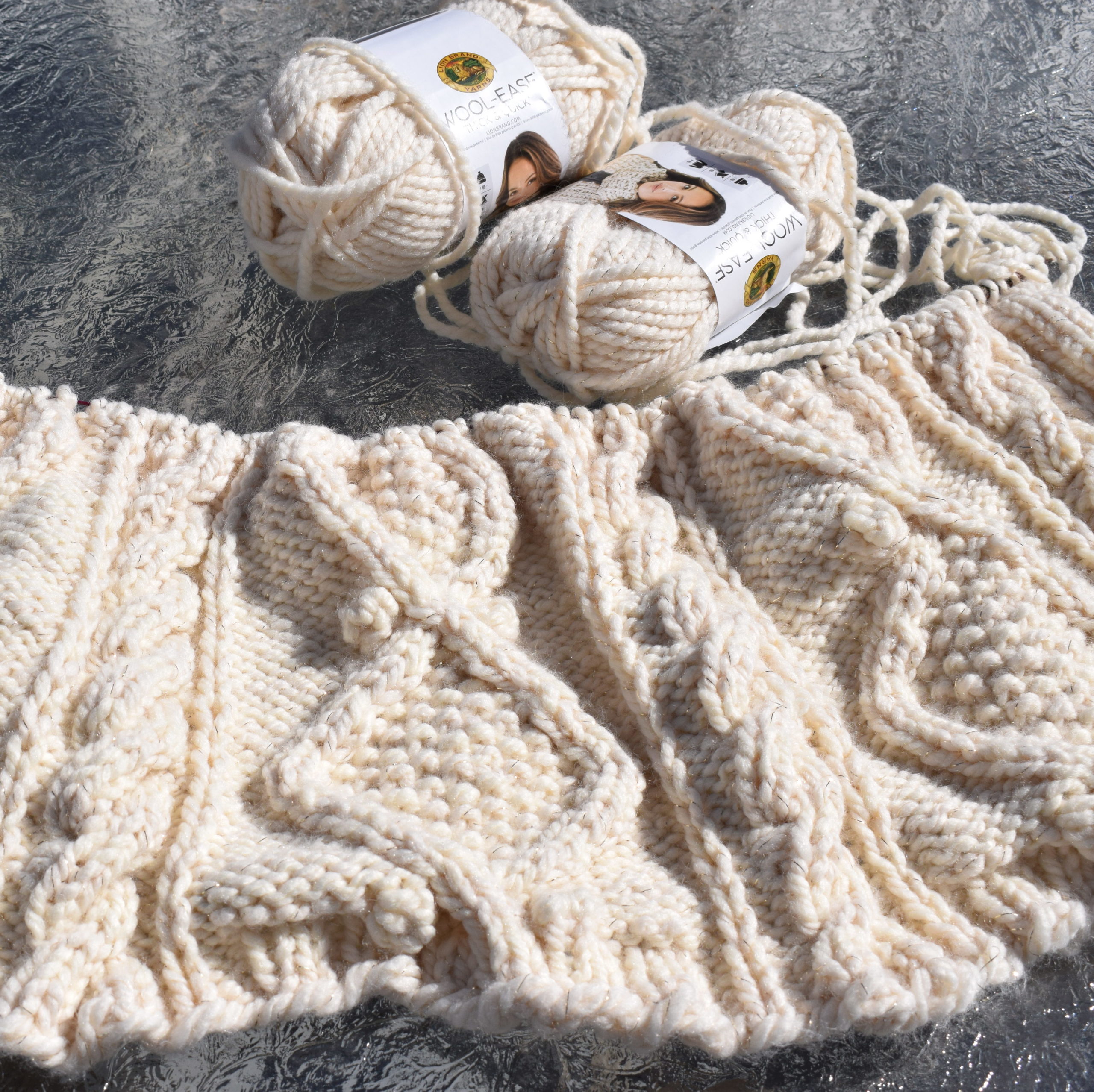 A partially-completed, deeply textured afghan knit in a bulky cream yarn.  Squishy cables bracket stitches forming large diamonds with seed stitch centers.  Two skeins of yarn are lying next to the work.