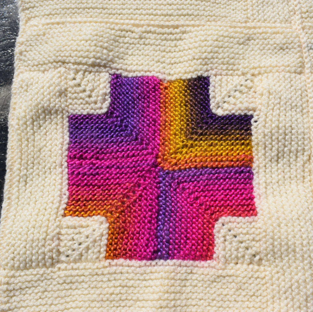 A blanket square of a mitered cross knit in a variegated yarn with purple, pink, orange, yellow, and navy stripes.  The cross is bordered by a log cabin frame in a cream colored yarn.