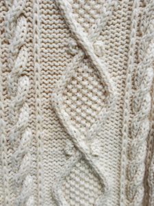 A close-up of the completed afghan, showing the seed stitch diamonds and bordering cable twists, worked in a cream colored yarn.