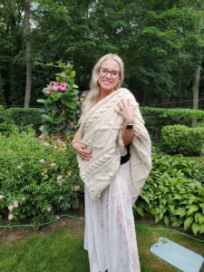 Alecia, the writer's cousin, poses in a garden wearing a white lace dress and black glasses.  She has draped her new white cabled afghan over one shoulder and is hugging it to herself.