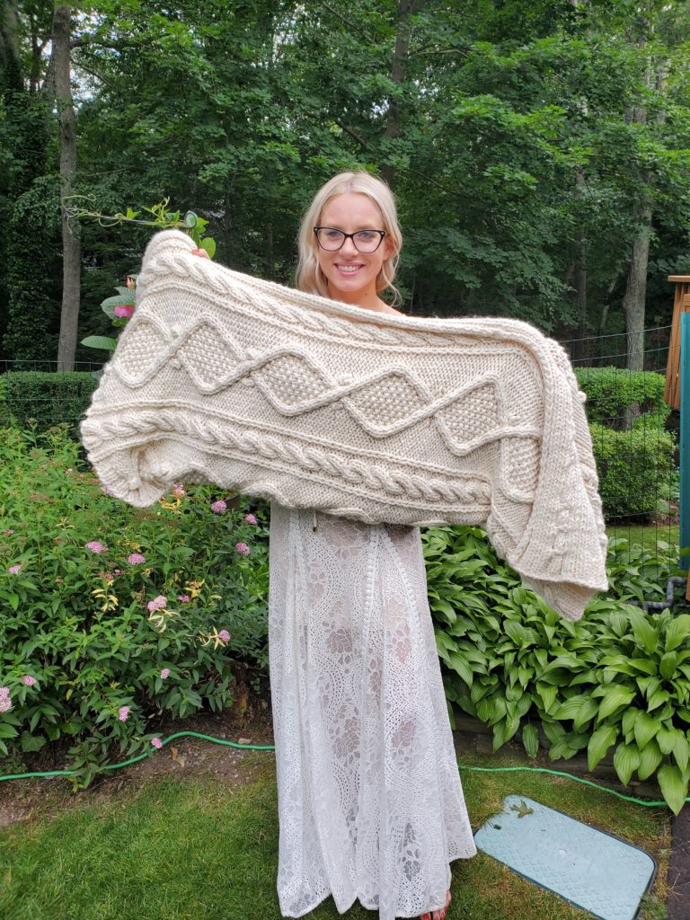 Alecia, the writer's cousin, poses in a garden wearing a white lace dress and black glasses.  She is holding out her new white cabled afghan and smiling.