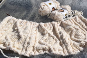 Blanket in progress is stretched across a table in the sunlight with two skeins of yarn laying next to it.